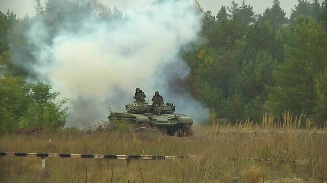 Two tanks with tankers at the top leave the forest and walk through the field. A large-caliber flame bursts from the muzzle. Real war scene with heavy armored vehicles in combat.