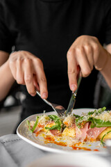 Close-up of a person cutting into a savory dish with arugula, ham, and grated cheese on a white plate, using a fork and knife. Concept of dining, homemade food, or restaurant meal.