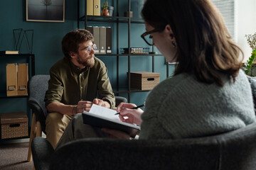 Man sitting in therapy session listening to woman therapist writing notes in notebook during...