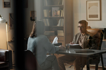 Man therapist listening to woman patient during therapy session in office, man holding clipboard...