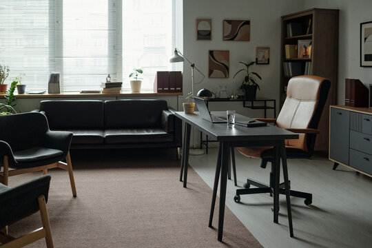 Empty modern therapy office showing desk with laptop and glass of water, armchair positioned for therapist, couch and chairs arranged for client sessions, large window providing natural light