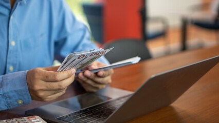 Person counting cash while reviewing documents and using a laptop, representing financial control,...
