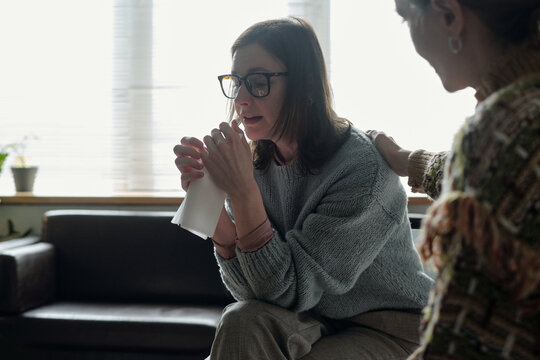 Woman sitting and holding tissue while crying, receiving comforting gesture from another woman during therapy session in office setting - Powered by Adobe