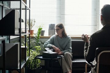 Woman sitting on sofa engaging in therapy session with man gesturing while talking, indoor office...