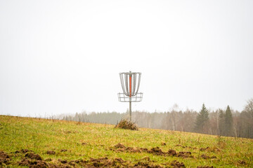 Disk golf basket (Frisbee golf) on grass field hill with forest in the background on a bright day cloudy sky background