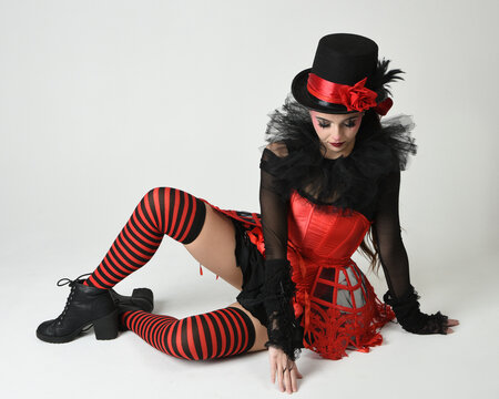 Burlesque circus performer, fantasy female model in halloween jester clown costume with top hat, red gothic corset. theatrical sitting pose kneeling on floor, isolated white studio background.