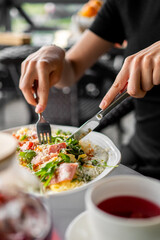 Close-up of a person eating a colorful meal with ham and greens using a fork and knife, served on a white plate with a red drink beside it, outdoor cafe setting.
