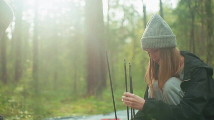 Couple preparing tent in tranquil forest as woman aligns multiple tent poles with focus, warm sunlight filters through trees creating gentle glow and peaceful camping atmosphere around them