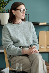 Portrait of woman sitting with hands clasped during therapy session, wearing glasses and looking...