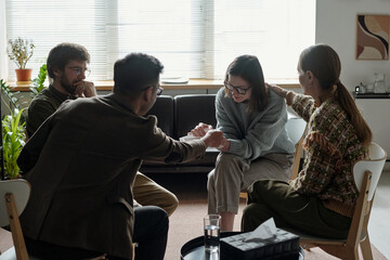 Multiethnic group of young adults sitting in therapy session, woman appearing emotional while Black...