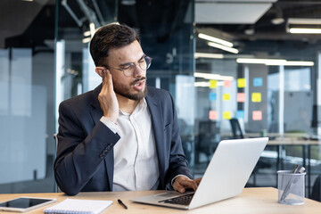 A man in a suit, experiencing ear pain while working on a laptop in an office setting