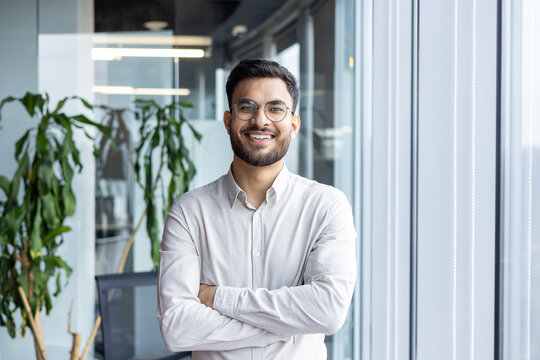 A smiling man with glasses stands confidently in a modern office setting, arms crossed.