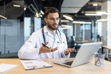 A doctor in a white coat uses a laptop for a video call, consulting or telemedecine, in a modern...