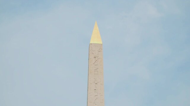 Upper part of Luxor obelisk in Paris. Tilt shot of a tall stone obelisk with hieroglyphs, set against a bright, cloudy sky. Close-up of the Luxor Obelisk with Egyptian hieroglyphs and golden top