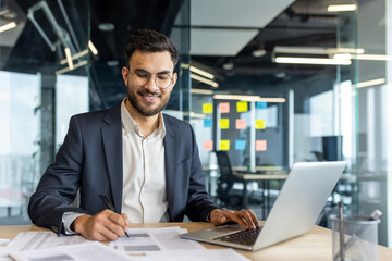 A smiling businessman in a modern office, working on a laptop while reviewing documents.