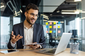 A businessman in glasses smiles while talking on a video call in a modern office setting.