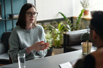 Woman sitting across from unseen engaging in therapy session, gesturing with hands while talking,...