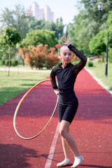 Focused young gymnast performing rhythmic jumps and poses with hoop on an outdoor track, showcasing...