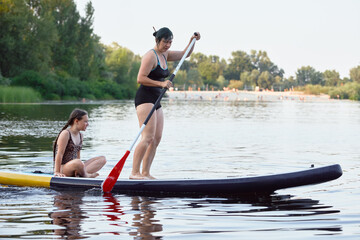 Mother and Daughter Enjoying Stand-Up Paddleboarding on a Calm River in Summer Daylight