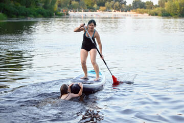A woman balances on a paddleboard as she assists a child in a calm lake setting, surrounded by...