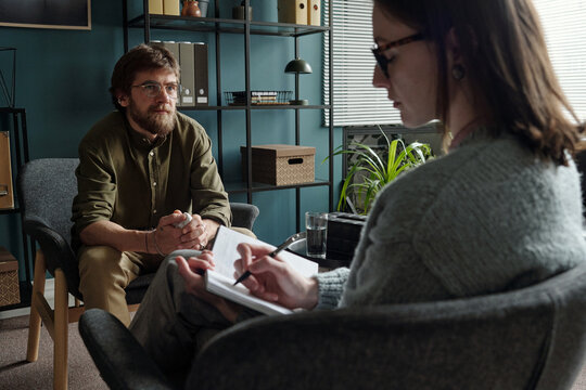 Man sitting in therapy session talking to woman therapist writing notes in notebook, both seated in modern office setting