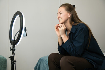 Young woman influencer is preparing for a live stream, adjusting ring light and smartphone in her...