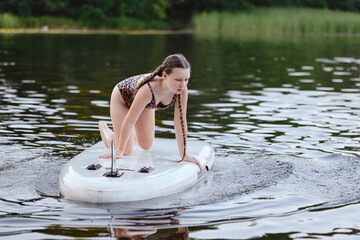 A young girl is kneeling on a paddleboard while enjoying a peaceful day on a calm lake. The...