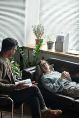 Woman lying on couch talking to man holding notebook during therapy session in office with plants...