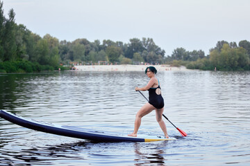 A woman is enjoying paddleboarding across a peaceful lake on a sunny day, surrounded by lush green...