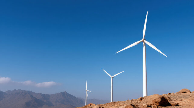 Three wind turbines stand on rocky terrain under a clear blue sky with mountains in the background, symbolizing renewable energy in a desert landscape.