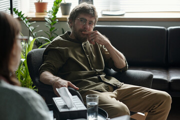 Man sitting in therapy session holding tissue box and looking thoughtfully at therapist, indoor...