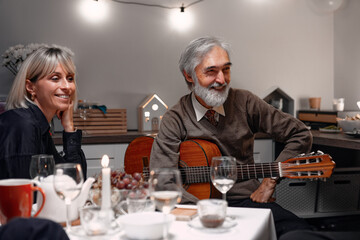 A cheerful elderly man plays the guitar while enjoying time with friends at a social dinner. The atmosphere is warm and inviting, with soft lights, decoration, and smiles filling the space with joy.