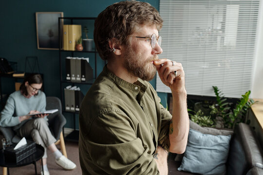 Man standing by window thinking with hand on chin while woman sitting in background writing on clipboard during therapy session