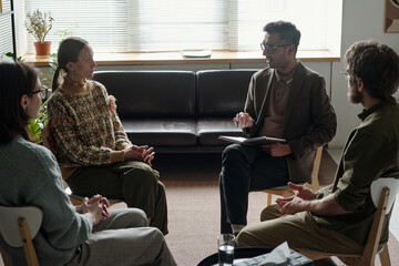 Group of multiethnic men and women sitting in therapy session, male therapist holding notebook and...