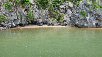 Limestone cliffs and lush greenery, with a small sandy beach and calm emerald sea of Thiên Cung cave, Ha Long Bay, Vietnam.