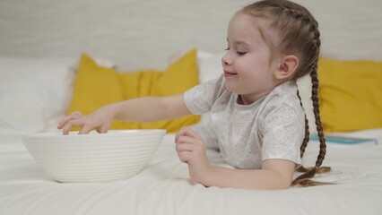 girl eats potato chips lying on bed and laughs, quick meal for kid, small child snacking on crispy potatoes, good appetite of cheerful child, fatty high-calorie fast food of carbohydrates, enjoy taste