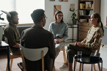 Group of and multiethnic men and women sitting in circle participating in therapy session,...