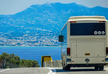 A white tour bus drives along a coastal road with a panoramic view of a seaside town and the...