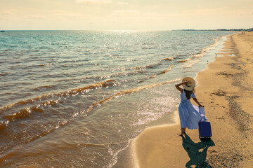 Young happy woman in a long pink dress stands on the beach with a travel suitcase in summer. The...