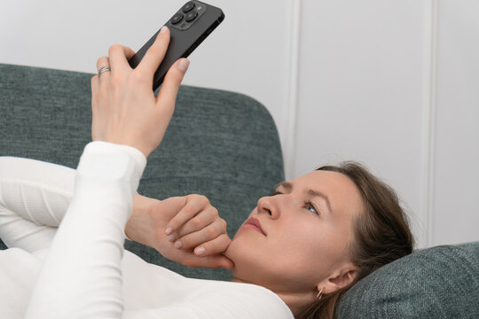 Young woman wearing a smartwatch is lying on a comfortable sofa at home, holding a smartphone above her and thinking with her finger on her lips