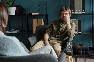 Man sitting in therapy session listening attentively to unseen therapist, resting chin on hand,...