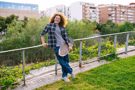 Portrait of a cheerful young man with curly hair holding his longboard and smiling in a city park