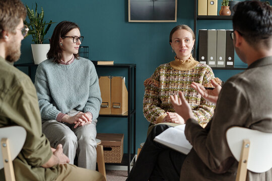 Group of multiethnic men and women sitting in circle participating in therapy session with professional therapist leading discussion holding clipboard