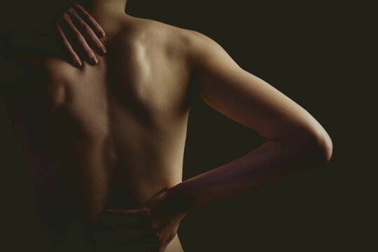 Woman's bare back and arms being lit by directional lighting in moody portrait on dark backdrop