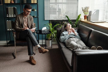 Man sitting in chair taking notes while man lying on sofa talking during therapy session in office...