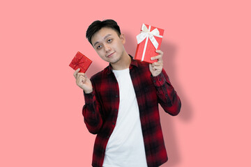 A young Asian man in a red plaid shirt and white t-shirt holds two red gift boxes, His facial...
