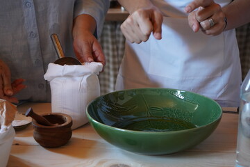 Two people preparing ingredients in a traditional kitchen setting. A green ceramic bowl with olive oil, a flour sack, and wooden tools highlight the authenticity of Mediterranean cooking.