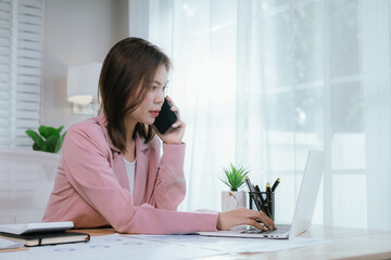 Businesswoman in pink coat working at desk using smartphone and laptop, managing tasks and planning projects in modern and organized office environment.