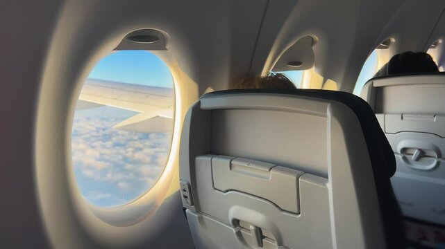 View on the fragment of the airplane cabin with passengers during the flight and a plane porthole through which you can see the wing of the plane and the blue sky with clouds in sunset.