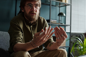 Man with beard and glasses sitting on couch gesturing with hands during therapy session, appearing...